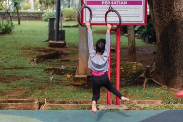 A little Asian girl hanging on ring pull-up bar outdoors in a public park with a sign showing the rules for using the equipment in Indonesian