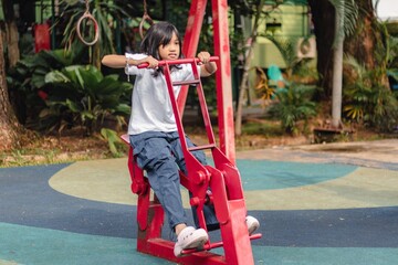 An Indonesian little girl trying an outdoor gym equipment in a city park. Iron exercise bike in a public park.