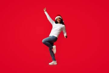 Fototapeta premium A joyful woman in casual clothing and a Christmas hat dances with excitement against a vibrant red background, celebrating the festive season with energy and happiness.