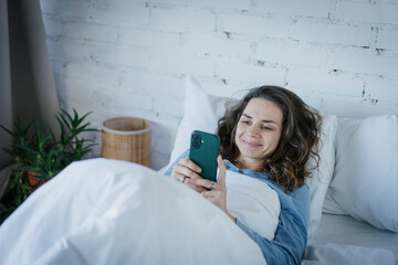 Morning Time. Smiling woman using smartphone browsing internet lying on bed at home