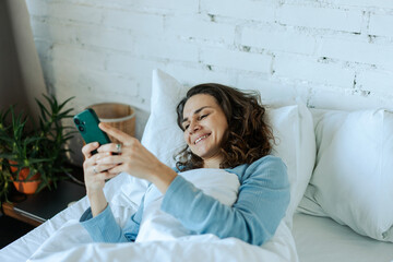 Morning Time. Smiling woman using smartphone browsing internet lying on bed at home