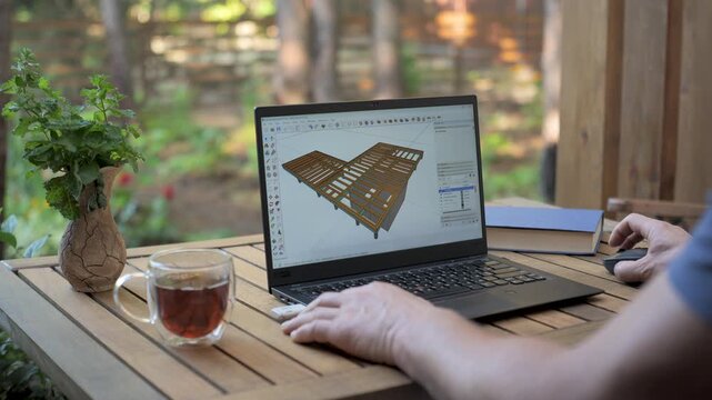 Architect working on a 3D model of a house on a terrace, a person using a laptop to design a 3D model of a house outdoors, with a cup of tea, a book and a plant on a wooden table