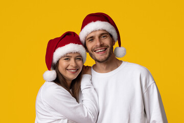 Fototapeta premium A cheerful couple smiles brightly while wearing matching Santa hats. They stand close together against a vibrant yellow backdrop, embodying the festive spirit of Christmas joy and togetherness.