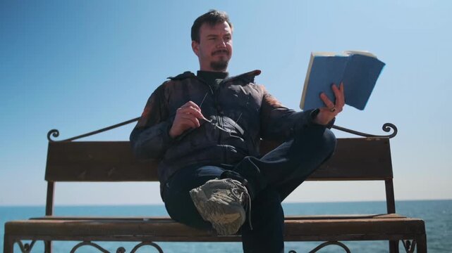 white man gesturing with book on bench by sea, expressive face, removing glasses, practicing monologue, lowangle composition, cross-legged pose, sunny sky, waterfront rehearsal, confident posture