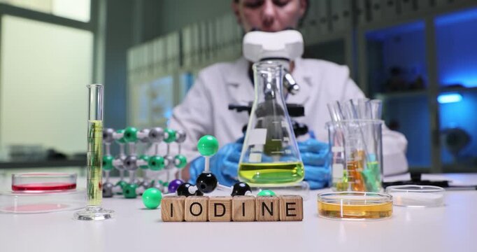Woman chemist studies iodine under modern microscope carefully. Miniature wooden cubes on table create word Iodine by laboratory beakers and molecular