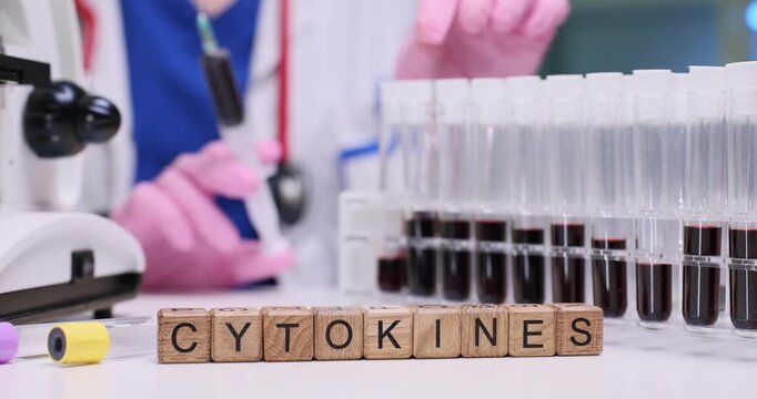 Doctor holds syringe filled with blood and examines test tubes prepared for analysis. Wooden cubes create word Cytokines highlighting focus of study