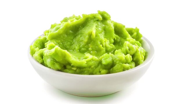 A closeup of a bowl of green mashed vegetables, possibly guacamole, on a white background. The bowl is white, and the vegetables are a vibrant green.