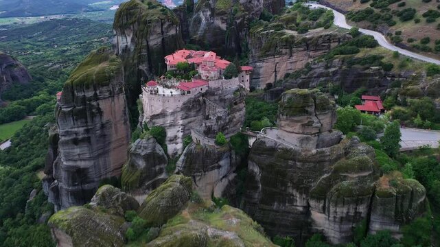 Aerial view of the Meteora Monastery complex perched atop towering rock formations, juxtaposed against verdant greenery and winding paths, Kalampaka, Trikala, Greece.