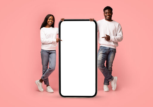 A happy millennial black couple stands in a pink studio, pointing at a big modern cellphone with an empty screen. They showcase their enthusiasm for a new mobile application together.