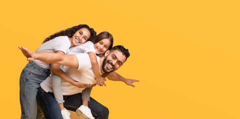 A joyful family is having fun in a vibrant yellow space, with two children playfully riding on their father's back, all wearing casual white outfits and smiling widely.