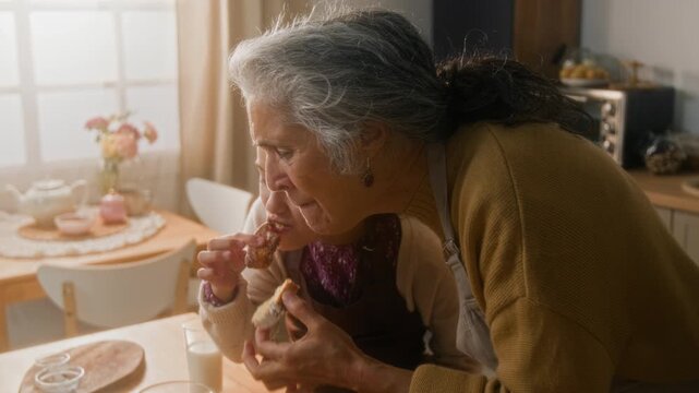 Tilt up shot of happy pre-teen girl and senior grandmother eating freshly baked loaf cake and high fiving at home kitchen