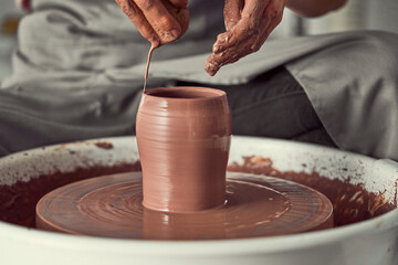 Brown-toned hands shape smooth clay on a spinning pottery wheel, forming a vase. Focused...