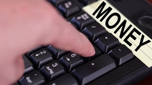 Un primer plano de un teclado con n&uacute;meros con el dedo de una mujer adulta marcando los n&uacute;meros y una nota amarilla con la palabra dinero