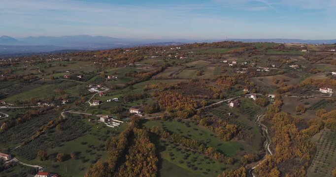 Aerial view of rolling hills dotted with sparse houses and trees displaying autumn colors under a clear sky, Gesualdo, Irpinia, Italy.