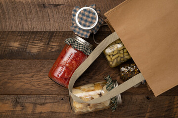 Food items preserved in glass jars spilling out of a brown, reusable farmer's market shopping bag onto a wooden table.