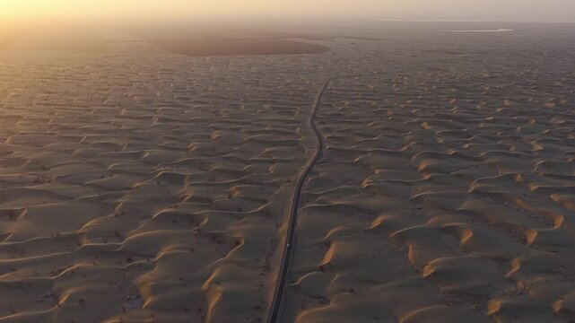 Aerial view of a road cutting through mesmerizing patterns of sand dunes, bathed in the warm glow of the desert sun, Taklamakan Desert, China.