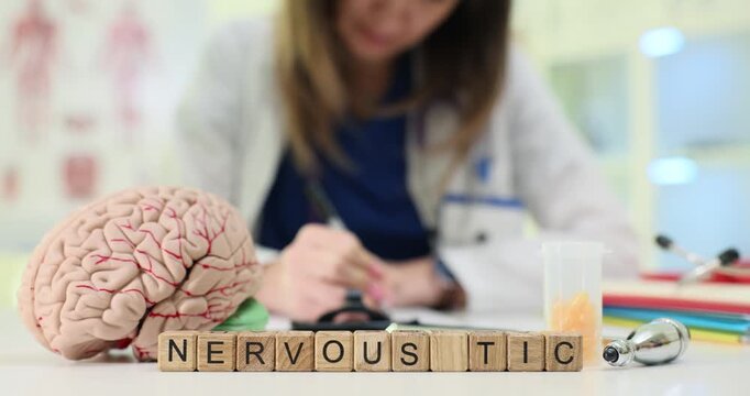 Wooden blocks spell word Nervous Tic near brain model on desk. Woman doctor carefully writes notes monitoring patient symptom history at appointment