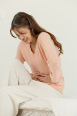 Naklejka premium Asian young woman abdominal pain holding stomach in bedroom wearing pink sweater and white pants concerned expression comforting herself with hand gesture
