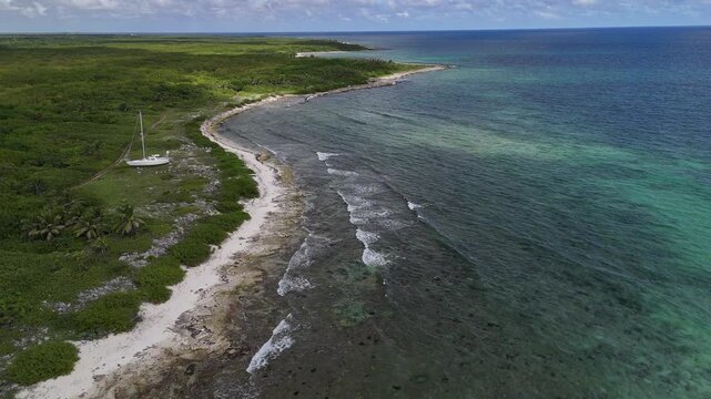 Aerial view of a serene coastline meeting the turquoise sea, next to a boat resting on the shore, a contrast of green vegetation and sandy beach, Mahahual, Quintana Roo, Mexico.