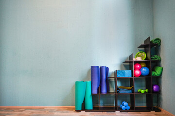 A wide shot of a gym corner with stacked yoga mats and a shelving unit of fitness equipment.