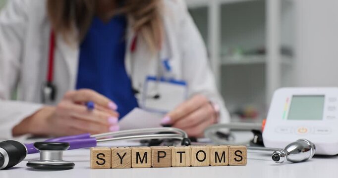 Wooden blocks spell word Symptoms near stethoscope and blood pressure monitor. Woman doctor writes chart noting pattern of disease signs in clinic
