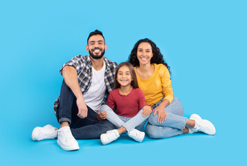 A happy family poses for a casual portrait in a studio. They sit on the floor, wearing colorful clothing, and share joyful expressions, showcasing a loving bond and warm atmosphere.
