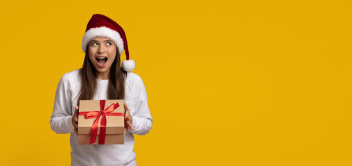 Obraz premium A woman wearing a festive Christmas hat appears surprised while holding a beautifully wrapped gift box with a red ribbon.