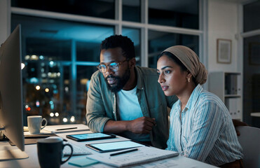 Diverse male and female colleagues focusing intently on a large computer monitor while working together in a modern office at night.
