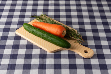 Fresh cucumber, carrot, and rosemary on a wooden cutting board over a blue checkered tablecloth. Healthy food, cooking, and preparation