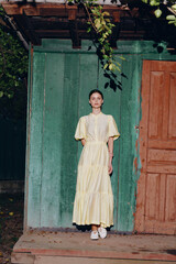 Naklejka premium Fashionable woman in a pale yellow dress stands before a weathered green wall and orange door, creating a serene rustic portrait with soft natural light.