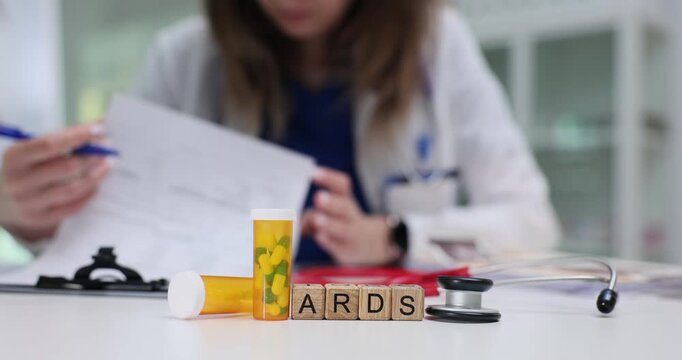 Doctor writes therapeutic recommendations in blank form. Miniature wooden blocks forming abbreviation ARDS lie arranged on table at drug capsules