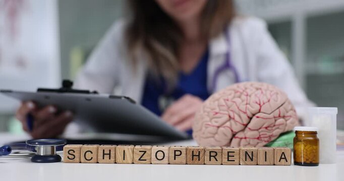 Woman doctor looks over documented episodes and medications. Wooden blocks forming Schizophrenia rest on desk near anatomical model of brain