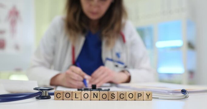 Woman doctor writes down recommendation for colonoscopy on medical form. Wooden blocks arranged on tidy examination table form word Colonoscopy