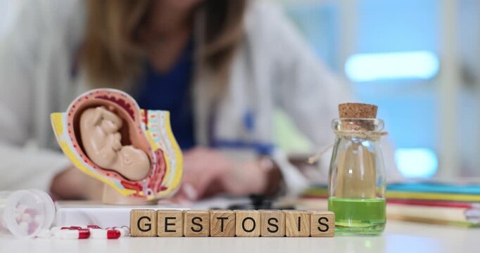 Wooden cubes forming word Gestosis rest on table near model of embryo in uterus. Doctor provides guidance on managing pregnancy complications
