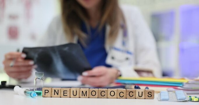 Woman physician analyzes lung radiograph with focused attention. Wooden cubes forming word Pneumococcus lie on table to indicate suspected diagnosis