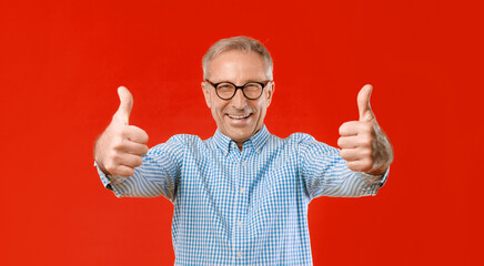 An older man stands with a big smile, holding up both thumbs in a sign of approval. He wears glasses and a checked shirt with a bright red background. His expression shows happiness. © Prostock-studio