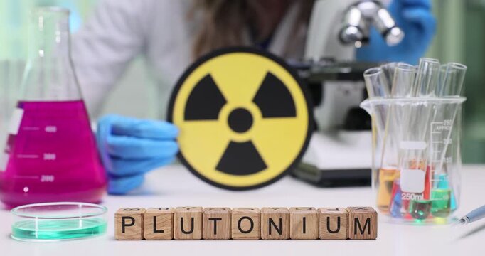 Laboratory worker shows warning sign indicating radiation danger. Wooden cubes form word Plutonium identifying subject of scientific analysis