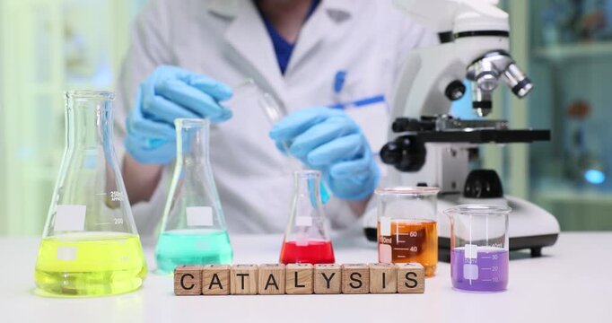 Laboratory worker pours azure solution into clean glass test tube. Miniature wooden blocks forming word Catalysis mark ongoing experiment in lab