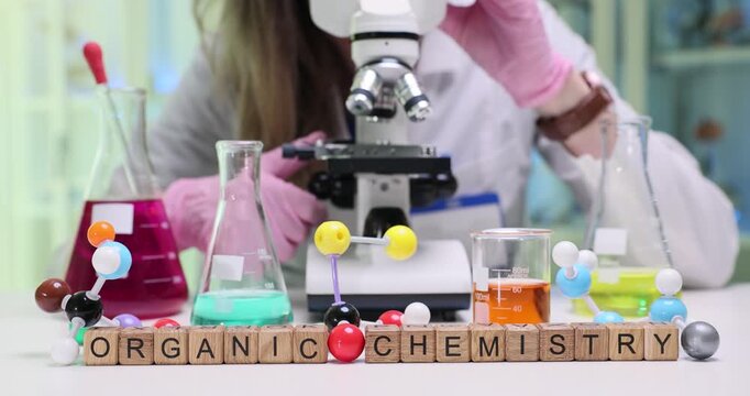 Woman researcher studies sample using modern microscope. Wooden cubes on table form phrase Organic Chemistry highlighting focus of laboratory study