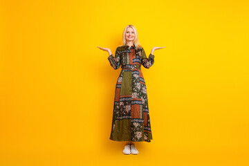 Elegant mature woman in patchwork dress stands against a bright yellow background smiling with open arms