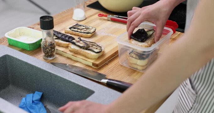 Housewife arranges sandwiches with fried eggplant in neat rows in container on countertop. Young woman stores sandwiches for later eating at lunch