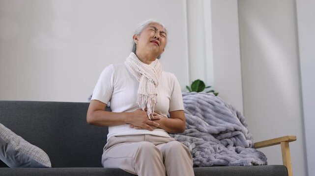 Elderly Asian woman suffering from stomach ache, feeling unwell and sick with a painful expression from indigestion or gastritis.
