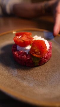 close-up of a female hand rotating a plate on a table containing a portion of meat tartare with cherry tomatoes and stracciatella