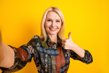 Smiling mature elegant woman in a patchwork dress gives thumbs up against a bright yellow background for fashion lifestyle and advertising photo