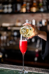Bartender pouring a vibrant, red cocktail into a tall champagne flute in a moody bar setting with blurred background and bokeh lights.
