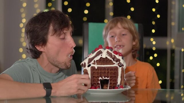 Joyful moments - father and son bite into the gingerbread house they decorated with fresh berries. Cozy winter evening, family fun, creativity, and festive bonding. Slowmotion video.