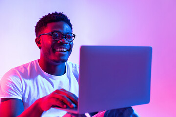 A young black man is smiling while using a laptop for online work in a colorful neon-lit environment. He is engaged in a remote meeting, showing enthusiasm and focus during the session.