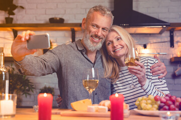 Adorable senior couple taking selfie on the phone during romantic date in the kitchen. Special event celebration. Spouses on videocall on Valentine`s Day. Dating concept