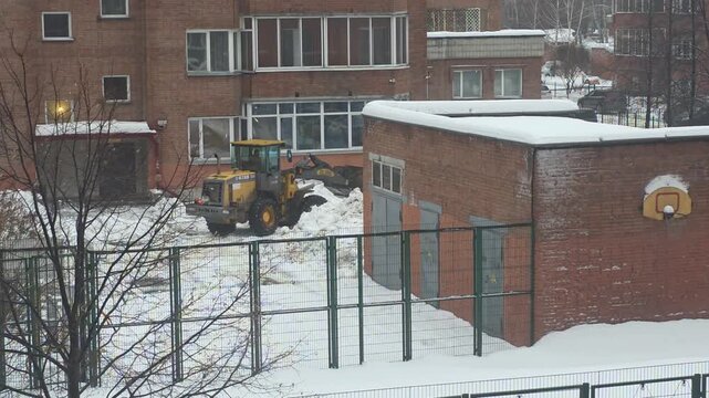 Yellow front loader clearing snow near a brick residential building and fenced playground in a snowy winter landscape
