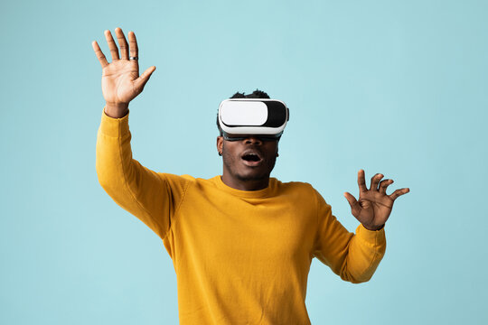 Emotional young african american man in yellow sweater using VR glasses, touching something invisible, millennial black guy experiencing virtual reality adventure on blue studio background, copy space - Powered by Adobe